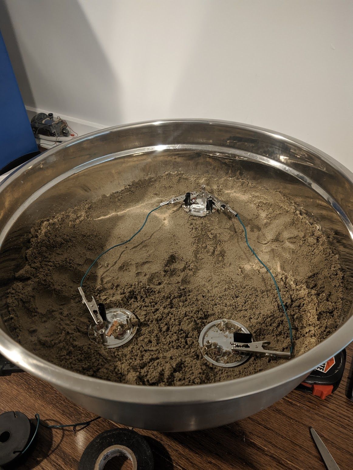 An aluminium pot half full with sand, with three kettle heating elements stuck inside